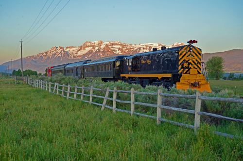 A vintage train travels through a green field with mountains in the background, under a clear sky at sunset.