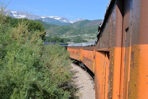 An orange train curves along a track surrounded by greenery and mountains under a clear blue sky.