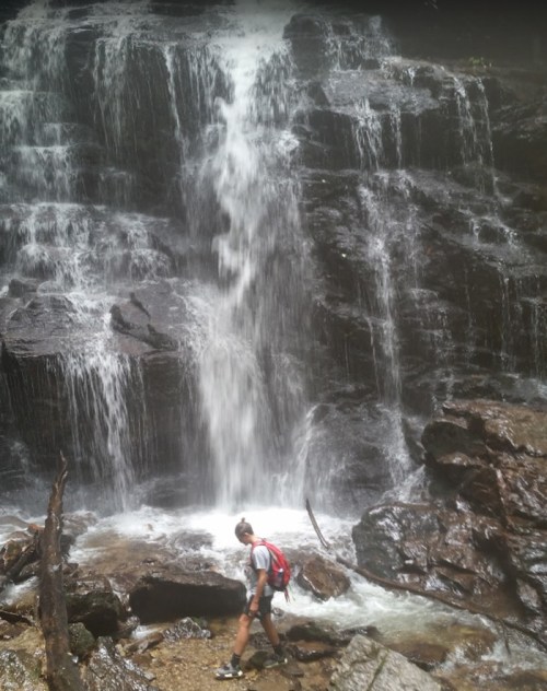 A hiker walks near a cascading waterfall surrounded by rocks and lush greenery.