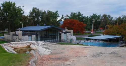 A modern building with a pool, surrounded by trees and a grassy area, under a cloudy sky.