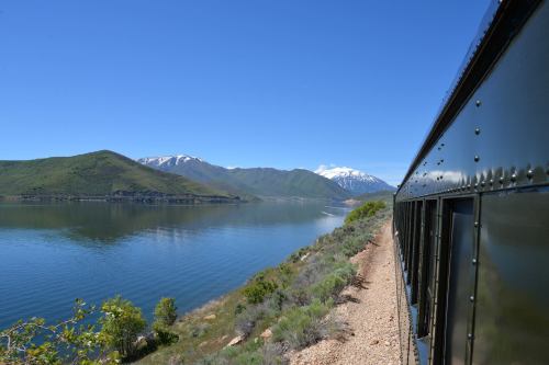 A scenic view from a train beside a lake, with mountains and snow-capped peaks in the background under a clear blue sky.