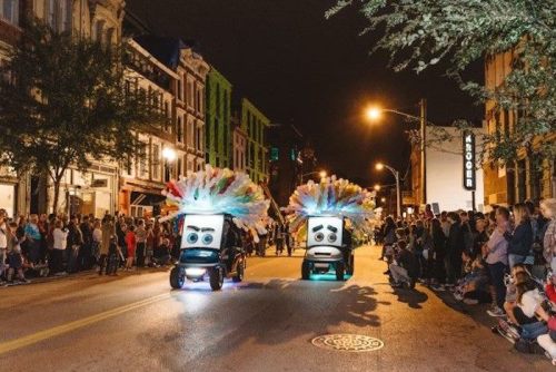 Two colorful, animated vehicles with feathered decorations parade down a street, surrounded by a cheering crowd at night.