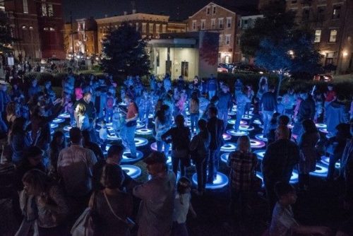 A vibrant night scene of a large crowd gathered around illuminated circular patterns on the ground.