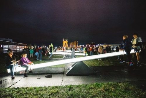People enjoying a nighttime playground with illuminated seesaws and a lively crowd in the background.