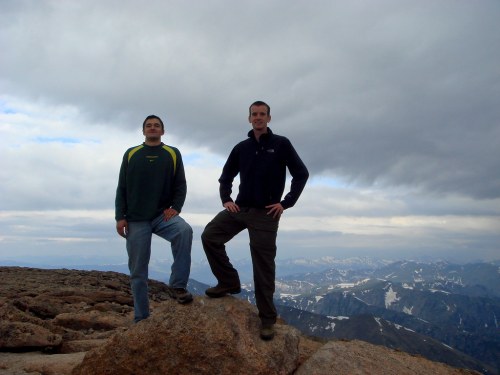 Two men stand on a rocky mountain peak, posing against a backdrop of cloudy skies and distant snow-capped mountains.