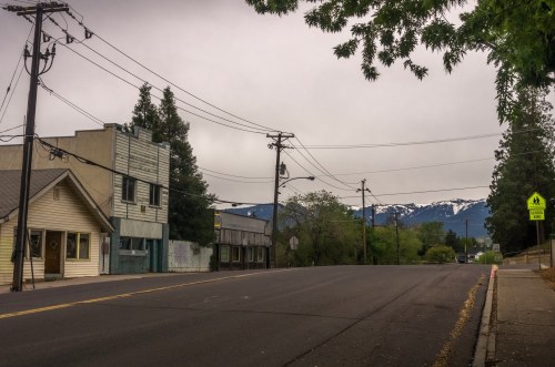 A quiet street scene with power lines, a few buildings, and mountains in the background under a cloudy sky.