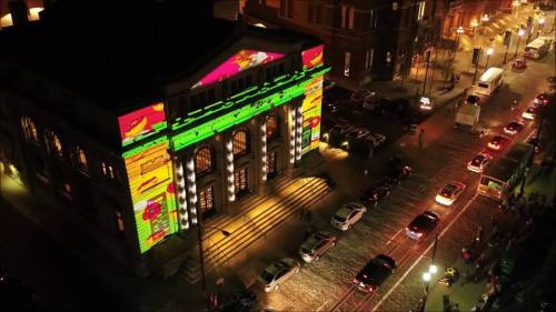 Aerial view of a building with colorful projections, surrounded by cars and people at night.