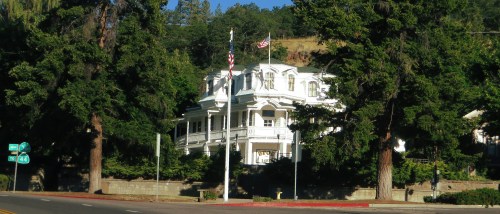 A large white Victorian-style house surrounded by tall trees, with flags flying in front and a road nearby.