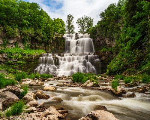A serene waterfall cascades over rocks, surrounded by lush greenery and a flowing river in a tranquil natural setting.