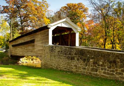 A wooden covered bridge spans a small stream, surrounded by colorful autumn trees and a stone wall.