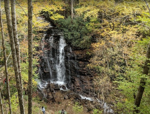 A serene waterfall cascades down rocky terrain, surrounded by vibrant autumn foliage and tall trees.