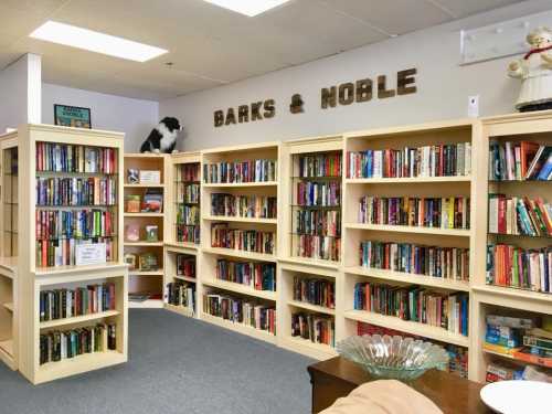 A cozy bookstore with wooden shelves filled with books, featuring a sign that reads "Barks & Noble" and a decorative dog.