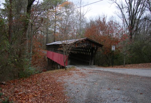 A rustic covered bridge with a red exterior, surrounded by autumn foliage and a gravel road.