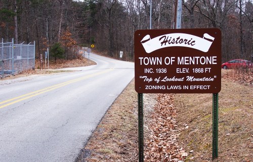 Sign for the historic Town of Mentone, indicating elevation and zoning laws, with a winding road in the background.