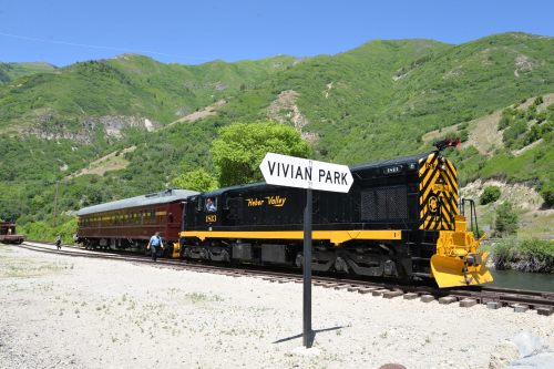 A vintage train at Vivian Park station, surrounded by green mountains and trees on a sunny day.