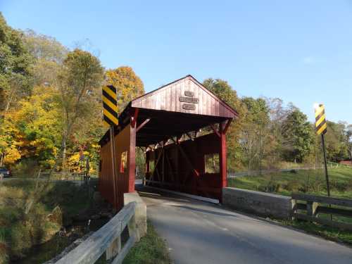 A red covered bridge spans a small creek, surrounded by autumn trees and clear blue skies.