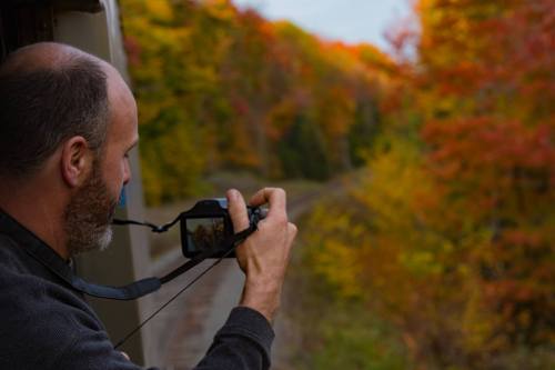 A man takes a photo of vibrant autumn foliage from a train window.