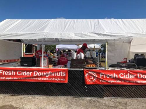 A food stall with a white tent, banners for fresh cider doughnuts, and people preparing doughnuts inside.