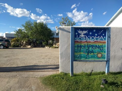Sign for La Paloma Hot Springs Spa, with a colorful design, surrounded by a gravel parking area and greenery.