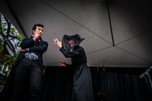 A man in a suit looks on as a woman in a black dress and veil gestures dramatically during a performance.