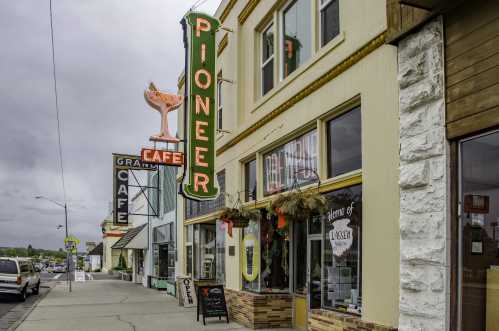 A street view of Pioneer Cafe with a vintage neon sign, surrounded by other shops and a cloudy sky.