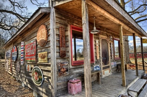 A rustic wooden building adorned with vintage signs and a porch, surrounded by trees and a cloudy sky.