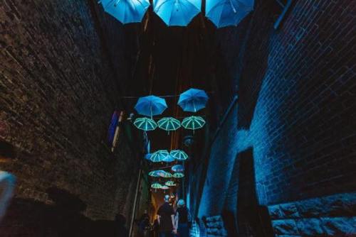 A narrow alley illuminated by blue light, featuring hanging umbrellas that create a whimsical atmosphere.