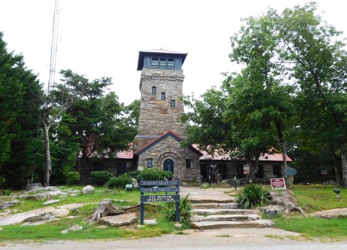 A stone building with a tower, surrounded by trees and rocks, featuring a sign in front.