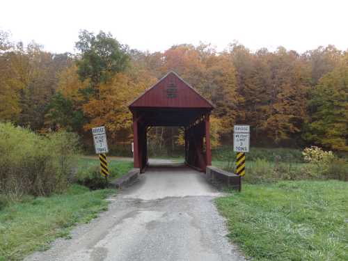 A red covered bridge spans a small road, surrounded by autumn trees with vibrant foliage. Signs indicate weight limits.