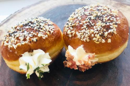 Two savory donuts topped with sesame seeds and poppy seeds, filled with creamy toppings, on a wooden surface.