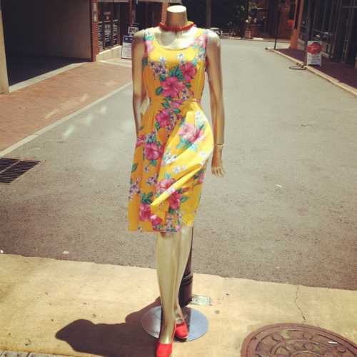 A mannequin wearing a yellow floral dress and red heels stands on a street, showcasing a vibrant summer outfit.