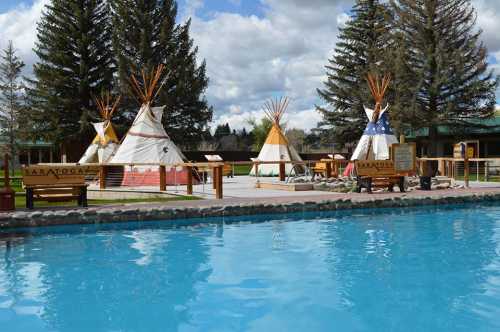 A serene outdoor scene featuring tipis by a swimming pool, surrounded by tall trees and a cloudy sky.