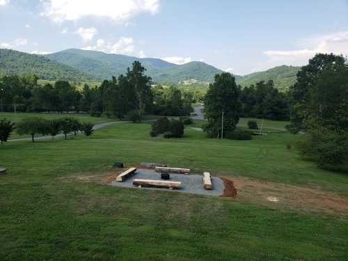 A scenic view of a grassy area with a fire pit surrounded by logs, framed by mountains and trees in the background.