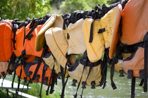 A row of colorful life jackets hanging on a rack near a river, with greenery in the background.