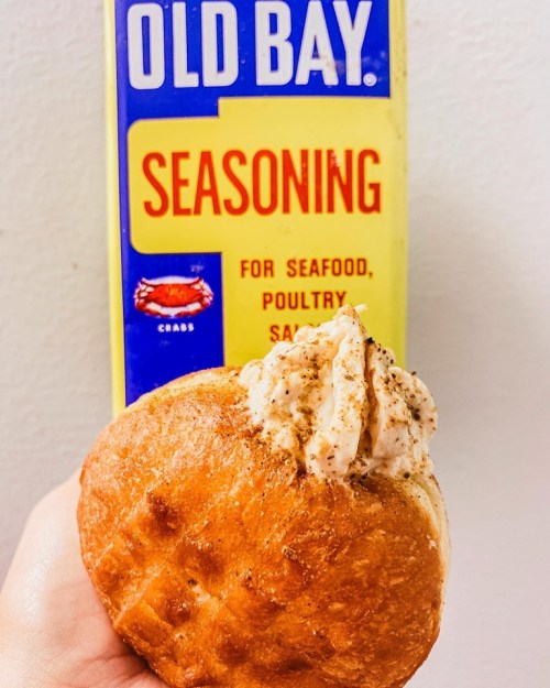 A hand holding a bun topped with creamy filling, with a box of Old Bay seasoning in the background.