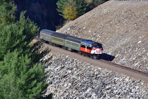 A vintage train with red and black engine and green passenger cars travels along a rocky hillside surrounded by trees.