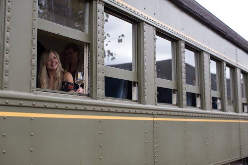 Two people smile from the windows of a vintage train, with one holding a drink.