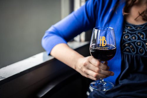 A person in a blue jacket holds a glass of red wine near a window, showcasing a logo on the glass.