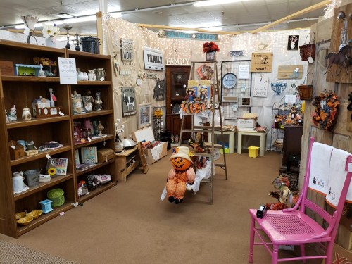 A cozy shop display featuring fall decorations, vintage items, and a scarecrow figure among shelves and wall art.