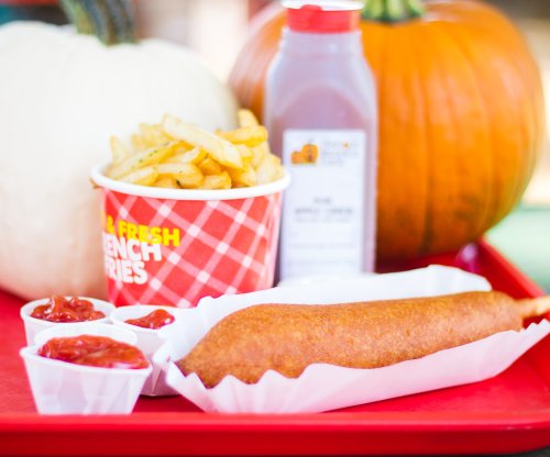 A tray with a corn dog, fries, ketchup cups, and a bottle of sauce, alongside a white pumpkin and an orange pumpkin.
