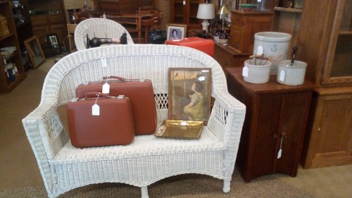 A white wicker loveseat with two brown suitcases, a framed picture, and decorative items on a nearby table.
