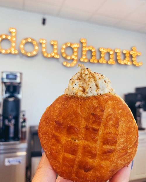 A hand holding a glazed doughnut topped with whipped cream, with a "doughnut" sign in the background.