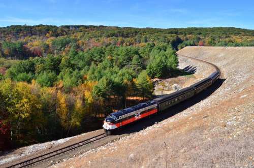 A colorful train curves around a hillside, surrounded by vibrant autumn foliage and clear blue skies.