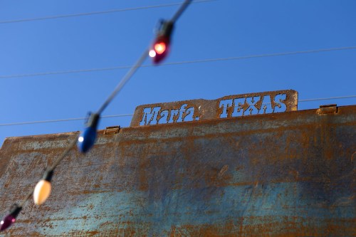 Sign reading "Marfa, Texas" on a rusted surface, with colorful lights in the foreground against a clear blue sky.
