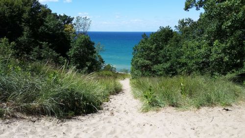 A sandy path leads down to a vibrant blue lake, surrounded by lush green trees and a clear sky.