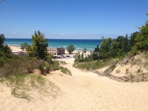 A sandy path leads down to a beach with clear blue water and trees under a bright blue sky.