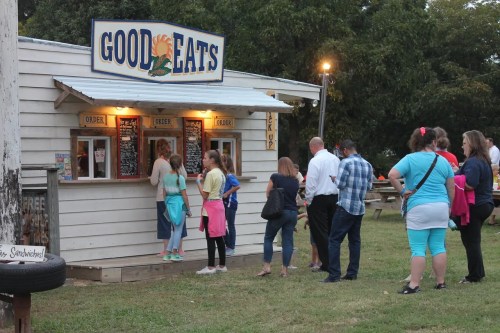 A group of people waiting in line at a food stand called "Good Eats" in a casual outdoor setting.