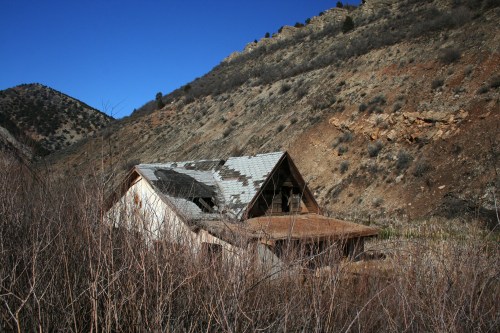 An abandoned house with a damaged roof, surrounded by dry brush and mountains under a clear blue sky.