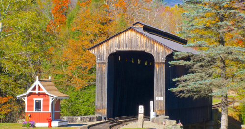 A rustic covered bridge surrounded by vibrant autumn foliage and a small red building nearby.