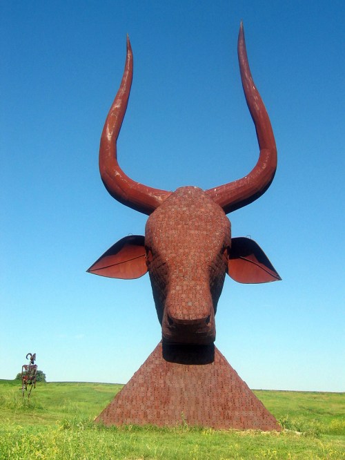 A large, rust-colored bull sculpture stands against a clear blue sky, surrounded by green grass.